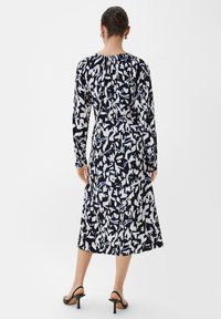 Schwarz-weißer gemusterter Langarmkleid mit fließender Silhouette. Verfügt über abstrakte Formen und einen gerafften Ausschnitt. Getragen mit schwarzen High Heels.