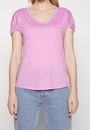 Femme portant un haut rose à manches courtes avec des manches froncées et un jean bleu clair, debout devant un fond uni et clair.