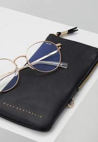 Pochette en cuir noir avec une fermeture éclair dorée, mettant en valeur un accent bleu. Lunettes de vue transparentes avec une monture dorée posées dessus. "QUAY AUSTRALIA" embossé.