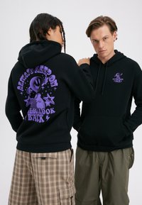 Dve osebi, ki nosita črne puloverje s kapuco; ena prikazuje zadnjo stran z vijoličnim tujcem in napisom "Vedno vztrajaj, nikoli ne glej nazaj", druga pa je obrnjena naprej.