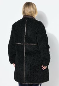 Cappotto in pile nero, con texture, dotato di colletto in pelle marrone, presenta un design a cuciture sul retro e una silhouette morbida.
