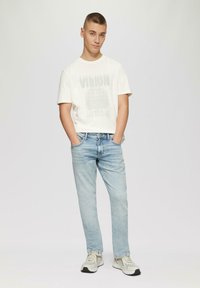 Witte graphic t-shirt met "VISION" print, lichtblauwe jeans en grijze sneakers. Gladde katoen kwaliteit, casual snit, handen in de zakken.