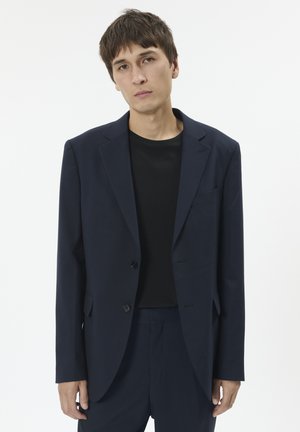 Marineblå jakkesætjakke med en struktureret, en- knaps design, med notch-lapel og to knapper; båret over en sort T-shirt.