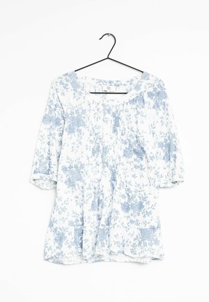 Blouse blanche avec motif floral bleu clair, manches trois-quarts, encolure ronde avec devant plissé, suspendue à un cintre noir sur fond blanc.
