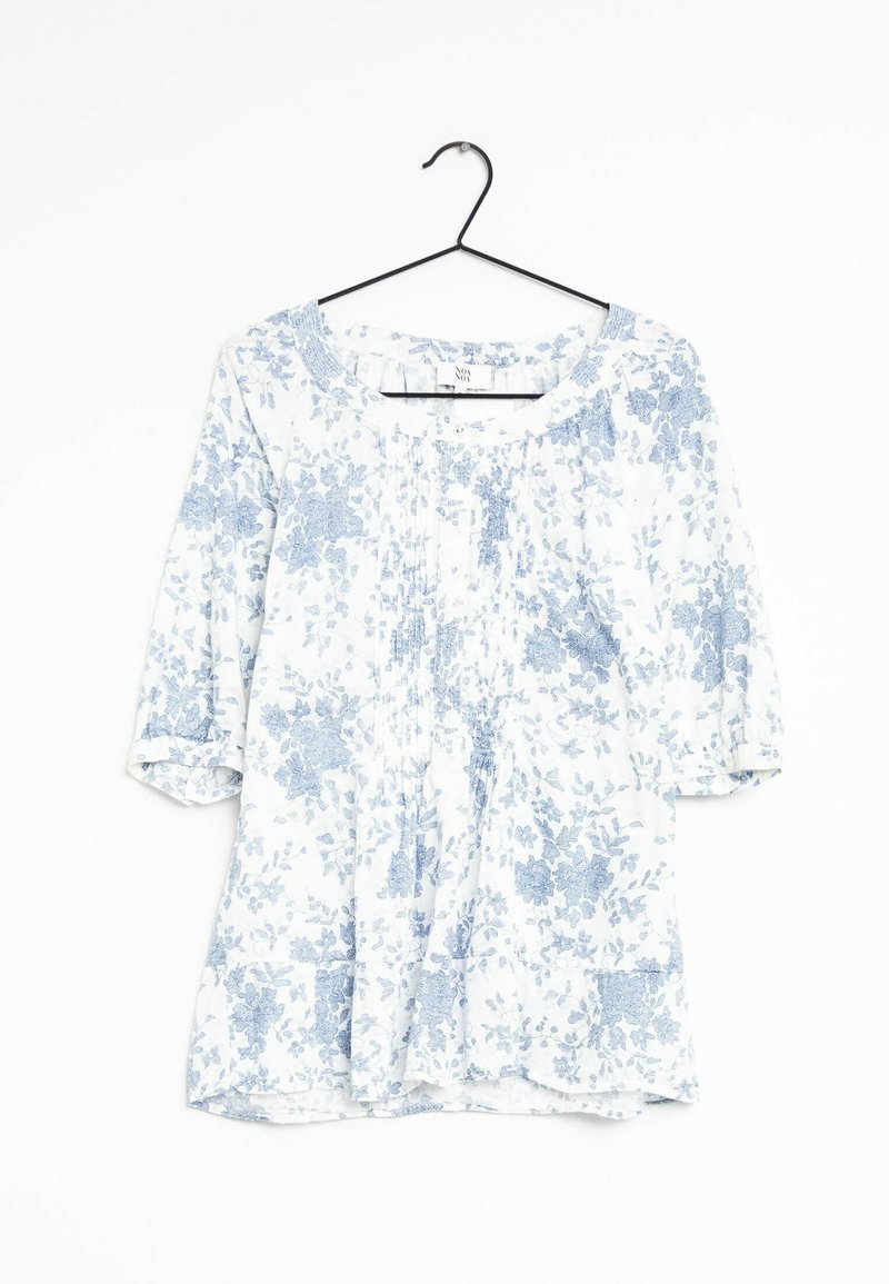 Blouse blanche avec motif floral bleu clair, manches trois-quarts, encolure ronde avec devant plissé, suspendue à un cintre noir sur fond blanc.