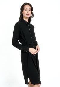 Must pikka, pika varrukaga nööbiga kleit kaelusega, kitseneva taljega ja külgsisselõigetega. Omab väikeseid valgeid nööpe ja sujuvat kangast tekstuuri.