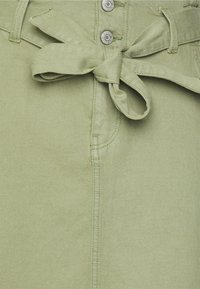 Tessuto verde chiaro con una texture morbida presenta una cintura legata, bottoni in metallo e dettagli di cucitura, mostrando un design ordinato.