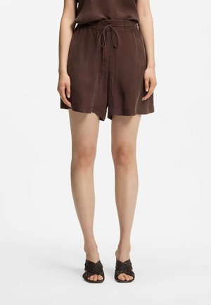 Nainen, joka on pukeutunut löyhiin ruskeisiin naruvyötäröisiin shortseihin ja vastaavaan yläosaan, yhdistettynä mustiin punottuihin avojalkaisiin liukusandaaleihin, seisomassa valkoisella taustalla.