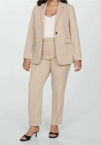 Costume beige sur mesure avec un blazer à un bouton et un pantalon slim, associé à un haut blanc côtelé et des talons noirs à bouts ouverts. Tissu lisse.