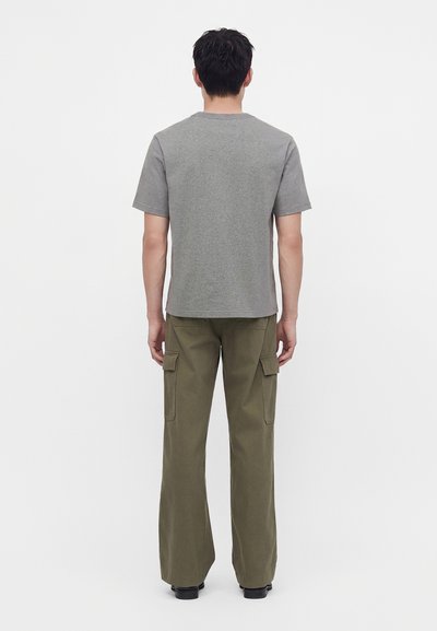 Homme de dos portant un t-shirt gris uni à manches courtes et un pantalon cargo vert olive avec des chaussures noires sur fond blanc.
