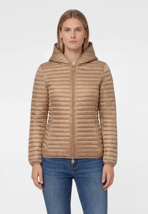 Donna con giacca trapuntata color cuoio con cappuccio e cerniera frontale, indossa jeans blu, in piedi contro uno sfondo chiaro e uniforme.