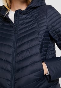 Chaqueta de plumas azul marino con capucha, presenta un cierre de cremallera y un patrón de acolchado horizontal. Textura suave y lisa con bolsillos laterales.