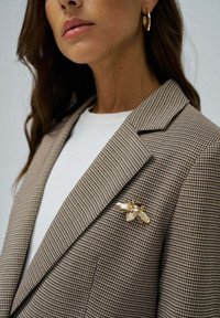 Blazer marrón de diseño houndstooth con solapas marcadas, llevado sobre una camisa blanca. Presenta un broche de abeja de resina dorada y transparente enganchado en la solapa.