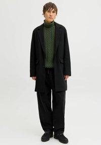 Manteau noir sur un col roulé en tricot vert, associé à un pantalon noir ample et des chaussures. Le design présente un motif texturé sur le pull.