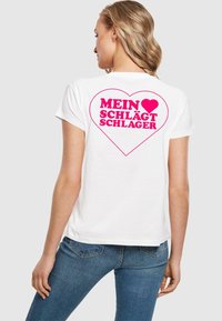 Balta medvilnė marškinėliai su dideliu rožiniu širdies piešiniu ir užrašu "MEIN ♥ SCHLÄGT SCHLAGER" ant nugaros. Trumpomis rankovėmis ir standartinio kirpimo.