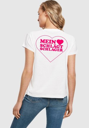 Valkoinen puuvillapaita, jossa on suuri vaaleanpunainen sydänkuvio ja teksti "MEIN ♥ SCHLÄGT SCHLAGER" selässä. Lyhyet hihat ja normaali istuvuus.