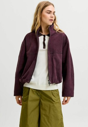 Femme portant une veste violet foncé ouverte, une chemise blanche et noire et un pantalon ample vert olive, debout devant un fond clair uni.