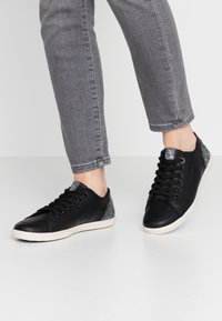 Baskets noires avec tige en cuir lisse, accents textiles gris sur le talon, semelle en caoutchouc blanche et lacets noirs. Adaptées pour un usage décontracté.