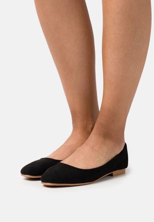 Bailarinas de ante negro con un diseño de punta redondeada y un tacón bajo en tono beige, sobre un fondo neutro. Textura suave y estilo minimalista.