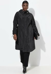Svart nylonjacka med huva, åtsittande midja och långa ärmar med texturerade detaljer. Bärs över svarta byxor och klackboots, håller en kuvertväska.