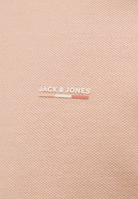 Artimas persikų spalvos tekstūruotos medžiagos vaizdas su "JACK & JONES" logotipu ir trimis horizontaliais spalvų juostelėmis po tekstu.