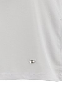 Weißes T-Shirt mit kurzen Ärmeln aus weichem Material, ausgestattet mit einem kleinen silbernen Metalllogo am unteren Saum und einer glatten Textur.