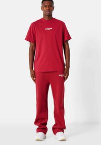 Rode katoenen T-shirt met korte mouwen en witte tekst, gecombineerd met bijpassende rode sweatpants. Witte sportschoenen met gele accenten maken de look af.