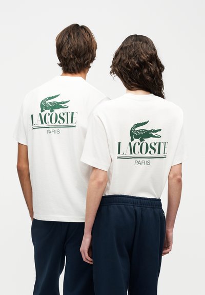 Двама души, облечени с бели тениски на Lacoste с лого на зелен крокодил и текст „LACOSTE PARIS“ на гърба, съчетани с тъмни панталони.