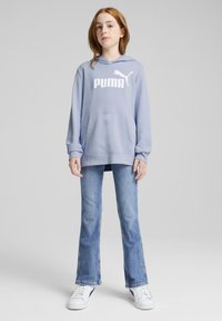 Svijetloplava dukserica s bijelim "PUMA" logotipom i kengurudžem džepom, uparena s proširenim plavim trapericama i bijelim tenisicama. Glatka tekstura tkanine.