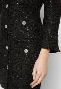 Gros plan d'une veste noire texturée avec des boutons décoratifs argentés et des sequins subtils, montrant la manche et une main reposant le long du corps.