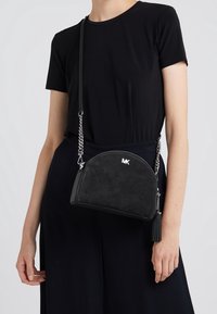 Svart mocka crossbody-väska med halvmåneform, featuring silverkedjeshalsband, logodetalj och en tofsaccent.