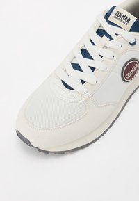 Vita och marinblå atletisk sko med nät- och mockamaterial, med rund logotypdetalj, platta snören och texturerad sula.