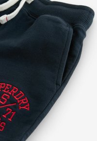 Mörkblå bomullsbyxor med struktur, dragsko i midjan och röd broderad logodetalj på låret.