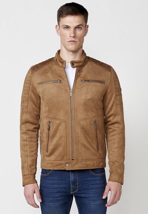 Chaqueta de ante color arena con cuello alzado, detalles acolchados en los hombros, bolsillos con cremallera en el frente y un diseño entallado. Se lleva con una camisa blanca y jeans.