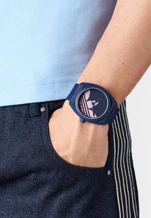 Sötétkék szilikon óra kerek számlappal, rózsaszín grafikai elemekkel. Az óraszíjak struktúráltak, a dizájn három óra mutatót tartalmaz.