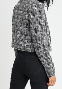 Veste courte en tweed noir et blanc avec un motif texturé, associée à un jean noir ajusté. Caractéristiques : manches longues et coupe décontractée.