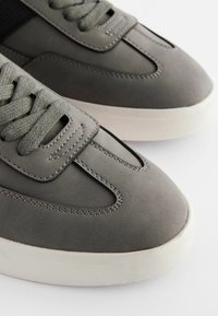 Sneakers grigi con una parte superiore in materiale sintetico liscio, lacci testurizzati, suola in gomma bianca e dettagli di cucitura sottili in tutto.