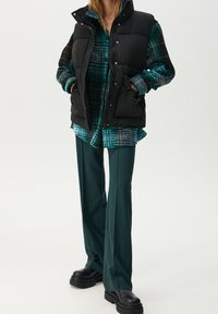 Personne portant un gilet matelassé noir, une chemise à carreaux verte, un pantalon vert foncé et des chaussures à plateformes noires, debout les mains dans les poches.