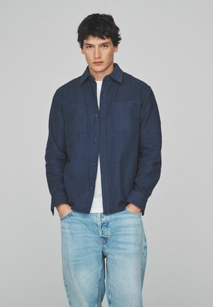 Junger Mann mit dunklen Haaren, der ein dunkelblaues Hemd über einem weißen T-Shirt und hellblauen Jeans trägt, steht mit den Händen in den Taschen.