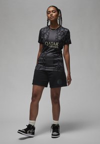 Musta urheilu-paita, jossa on leopardikuvio, varustettuna kultaisella "QATAR AIRWAYS" -logolla. Yhdistetty mustiin shortseihin ja korkeisiin mustiin lenkkareihin.