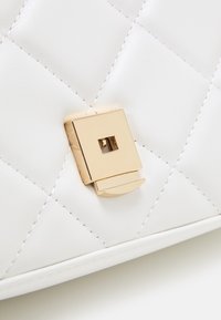 Vadderad vit läderhandväska med ett guld fyrkantigt spänne och en böjd detalj. Texturerad yta med synliga sömnadsdetaljer.