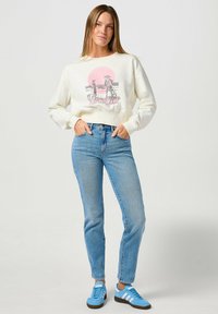 Sudadera crema con estampado gráfico de figuras y un sol rosa, combinada con jeans azul claros y zapatillas azules con acentos en blanco y negro.