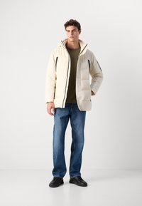 Gepolsterte Cremejacke mit hohem Kragen, schwarzen Akzenten und Reißverschlusstaschen, kombiniert mit blauen Jeans und schwarzen Schuhen.