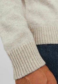 Jasno beżowy sweter wykonany z miękkiej dzianiny, z mankietami w prążki. Tekstura jest lekko puszysta, co komplementuje gładką powierzchnię.