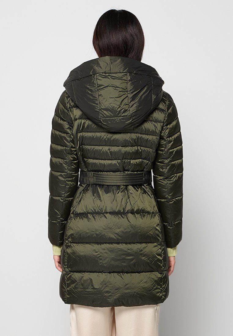 Frau in einem langen, olivgrünen, gesteppter Daunenjacke mit Kapuze und Gürtel, steht vor einem schlichten, hellgrauen Hintergrund.