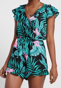 Zwarte romper met tropische bladeren en roze bloemenpatroon, korte ruches mouwen, tailleband en een ontspannen pasvorm. Soepele stofstructuur.