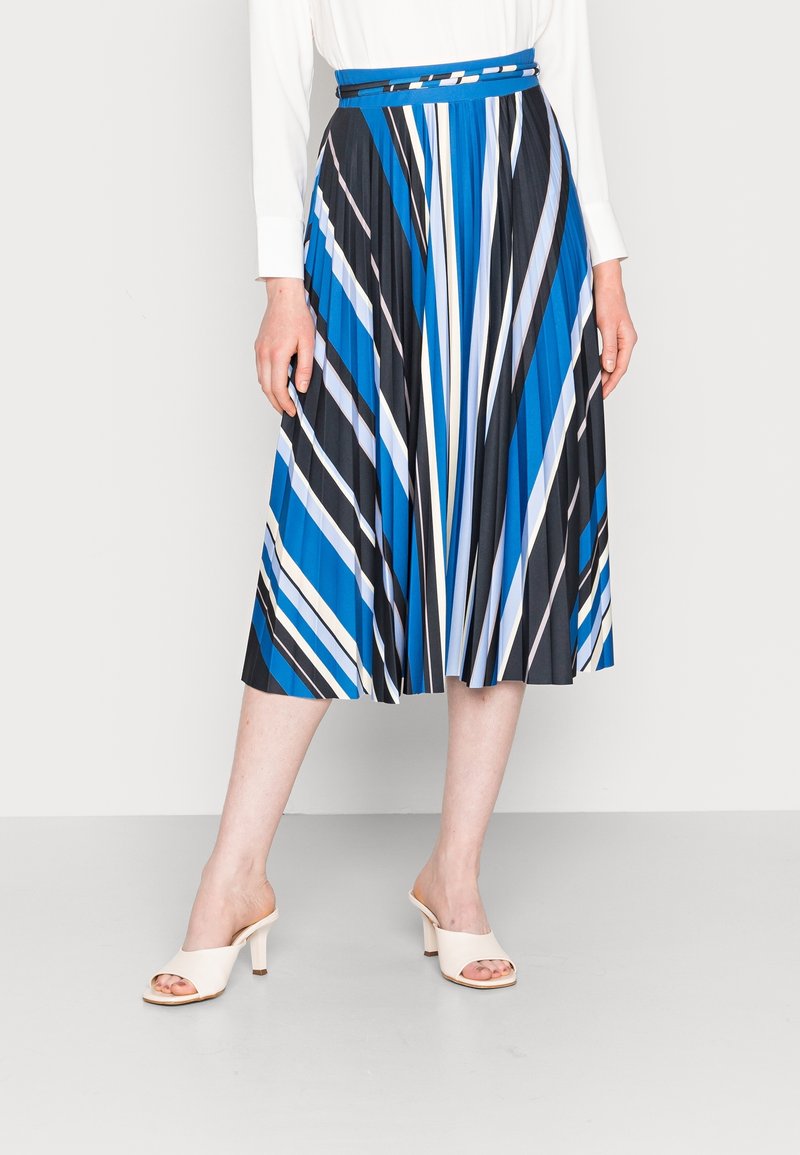 Femme portant une jupe midi plissée avec des rayures diagonales bleues, noires, blanches et grises et des sandales à talons blanches ouvertes au bout.