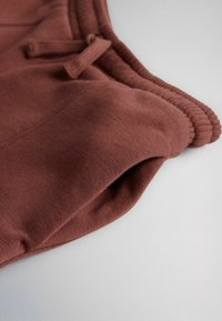 Rudos mezgimo sportinės kelnės su raišteliu liemens juostoje ir šonine kišene, išdėliotos plokščiai ant pilko paviršiaus.