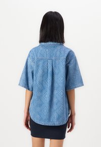 Camică din denim albastru cu mâneci scurte, având o textură subtilă cu modele, guler pliabil și un tiv rotunjit.