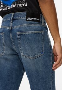 Mėlynos džinsinės kelnės su tiesiu sijonu, turinčios juosmens juostoje juodą prekės ženklo etiketę ir dvi galines kišenes. Audinys turi tekstūrinį paviršių.